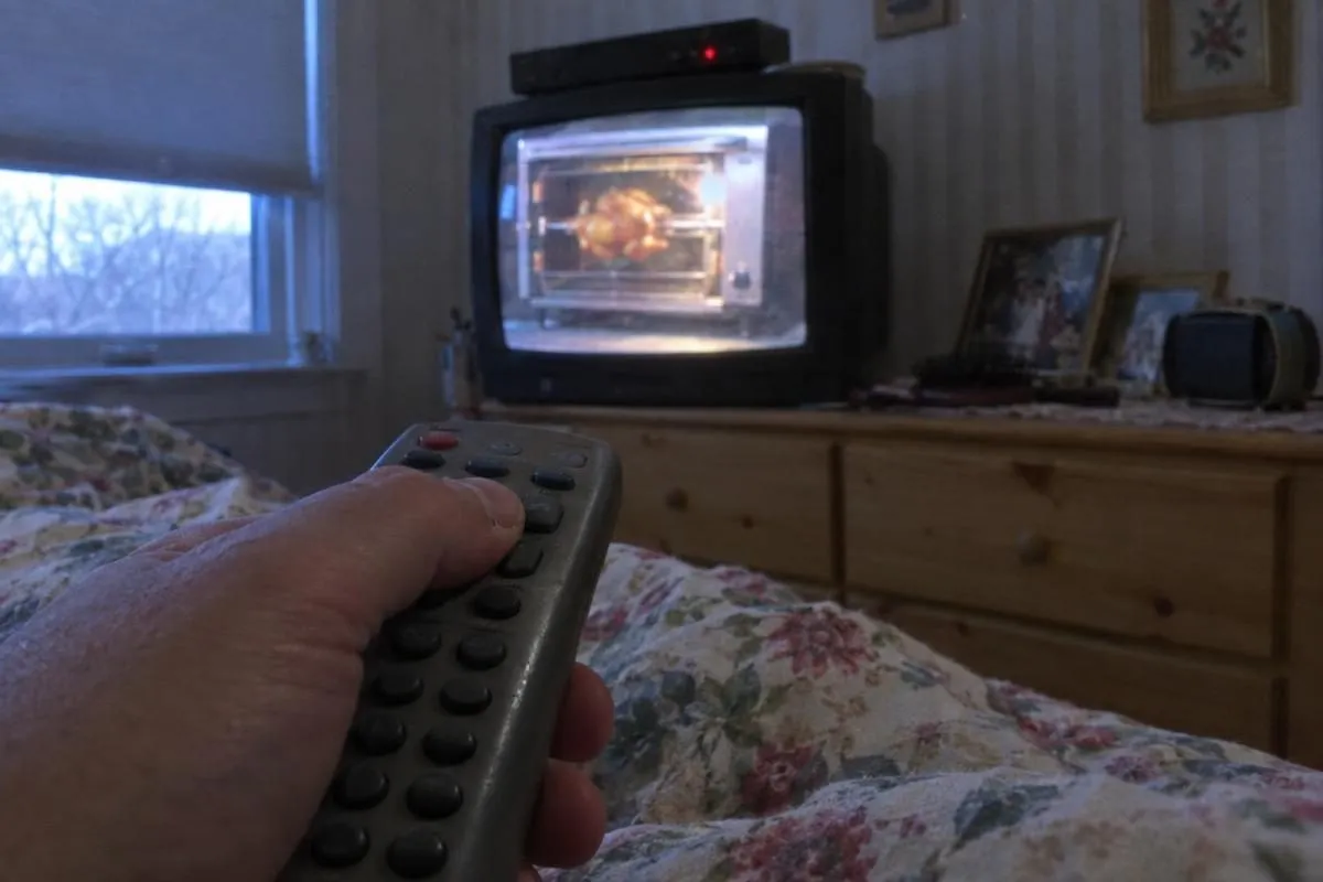 A hand holding a cable remote while a 90s infomercial for a rotisserie oven plays on a bedroom TV